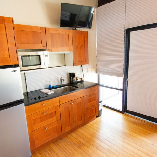 Small kitchen with wooden cabinets, stainless steel fridge, microwave, sink, coffee maker, television, and large window with blinds.