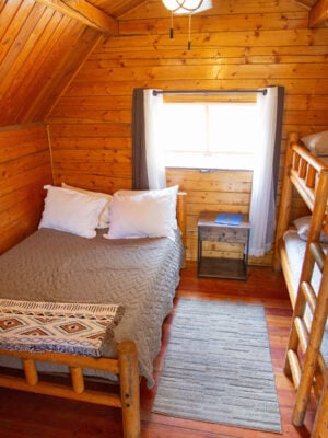 A cozy wooden cabin bedroom with a double bed, a bunk bed, a small nightstand, a window with curtains, and a rug on the floor.