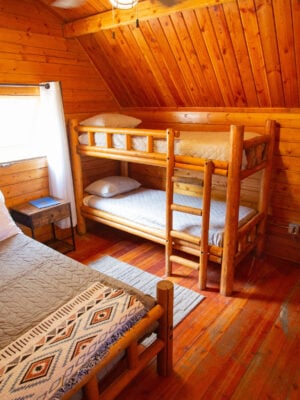 A wood-paneled room with a double bed, a bunk bed, a small black fridge with a microwave on top, a window with curtains, and a nightstand.