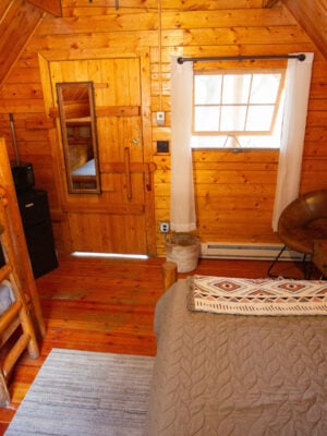 Small wooden cabin bedroom with bunk beds, a double bed, a chair, a bench, a mirror on the door, and a window with white curtains.