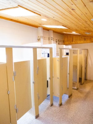 Public restroom with beige stalls, a long countertop with sinks and mirrors on the right, and a wooden ceiling.