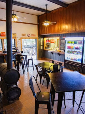 A small cafe or snack bar with tables, chairs, a wood stove, beverage coolers, and a counter; ceiling fans and windows provide light to the cozy interior.