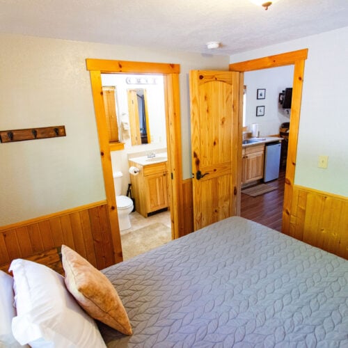 Small bedroom with a bed, wooden trim, an open door to a bathroom, and another open door leading to a kitchenette area.