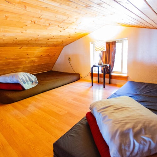 Small attic room with wood-paneled ceiling, two floor mattresses with pillows and blankets, a side table, lamp, and a window with curtains.