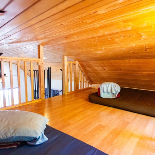 A loft with wooden walls and ceiling, two floor mattresses with pillows and blankets, and a railing overlooking the space below.