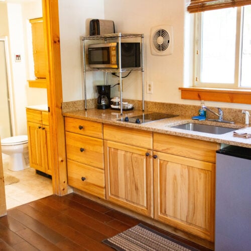 Small kitchen area with wooden cabinets, mini fridge, microwave, coffee maker, and sink; bathroom with toilet visible through open door on the left.