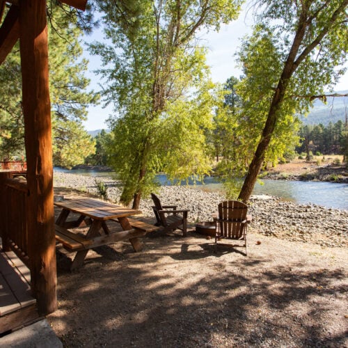 Wooden picnic table and chairs on a shaded riverbank next to a cabin, with trees and a flowing river in the background.