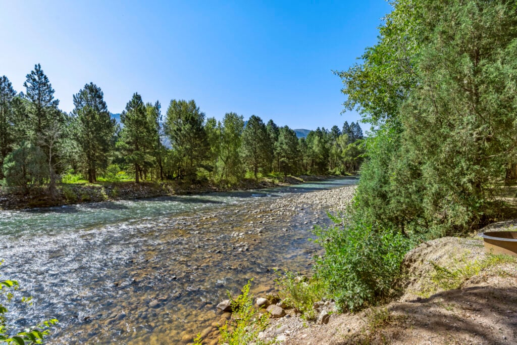 Clear river flows through a rocky, tree-lined landscape under a bright blue sky with sunlight illuminating the scene.