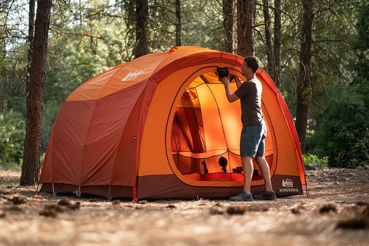 A person stands outside an orange REI tent in a wooded area, taking a photo with a camera. Sunlight filters through the trees onto the campsite.