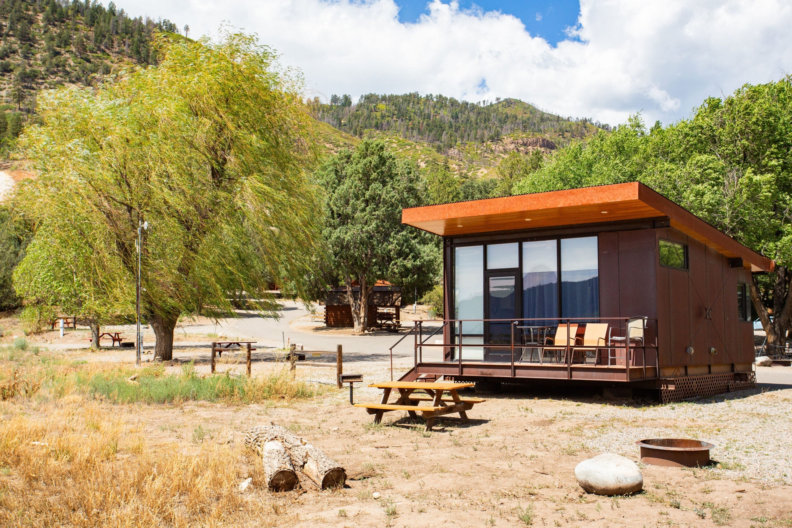 A modern tiny house with a patio sits in a dry, grassy area surrounded by trees, hills, a picnic table, and a fire pit under a partly cloudy sky.