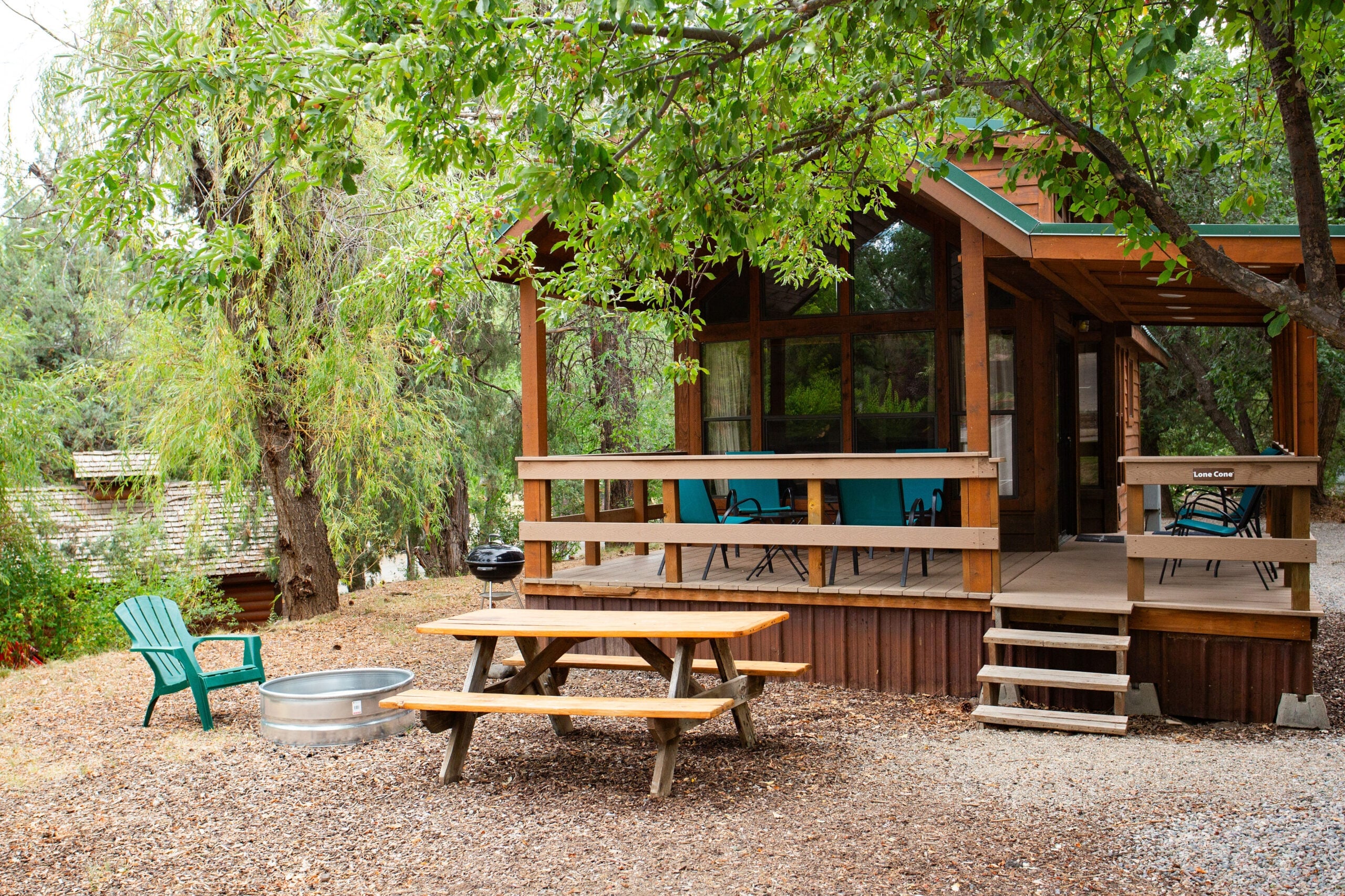 A wooden cabin with a covered porch, picnic table, fire pit, and green chair surrounded by trees and gravel in an outdoor setting.