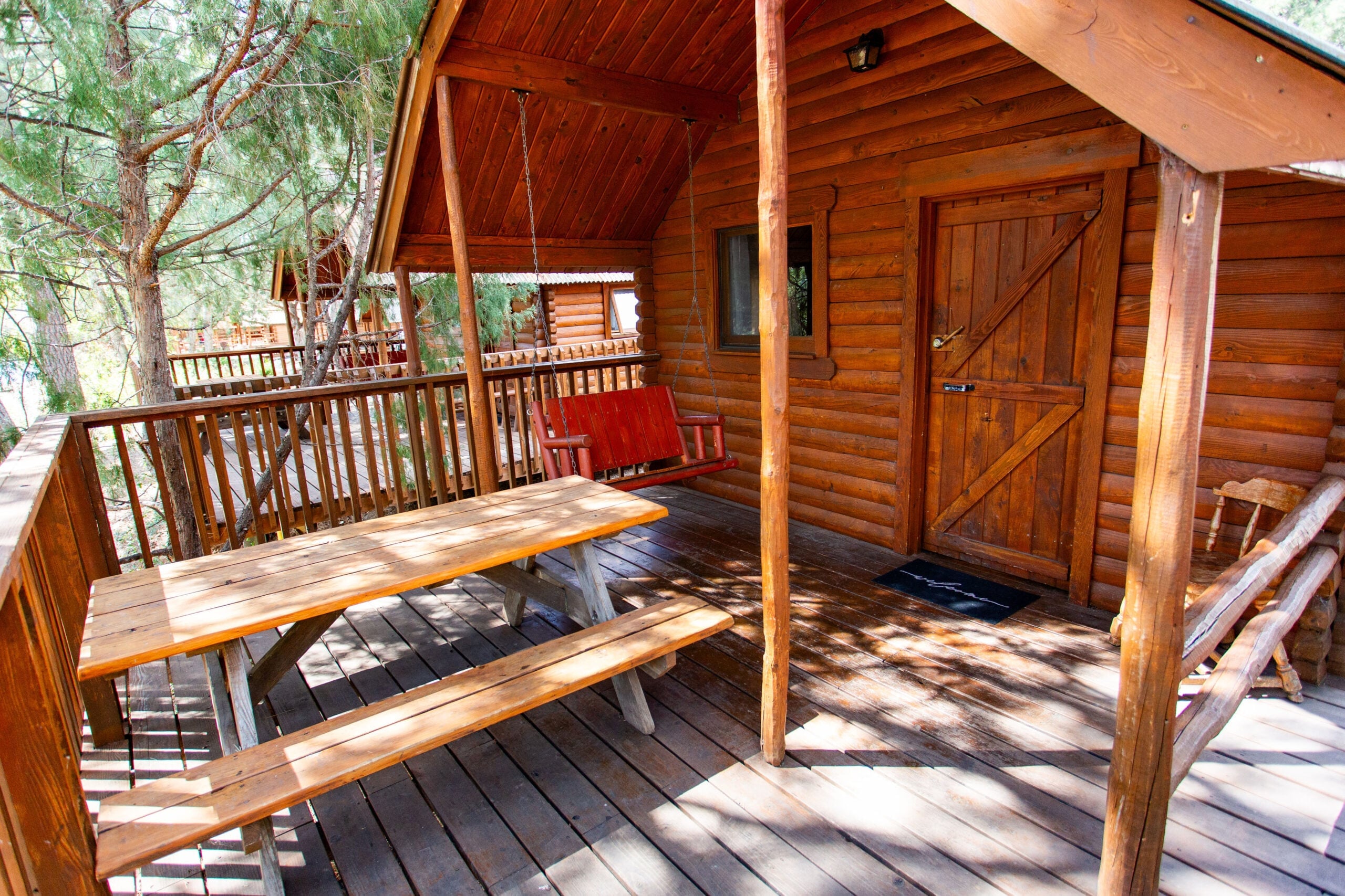 Wooden cabin porch with a picnic table, a red porch swing, and a wooden door, surrounded by trees and wooden railings.