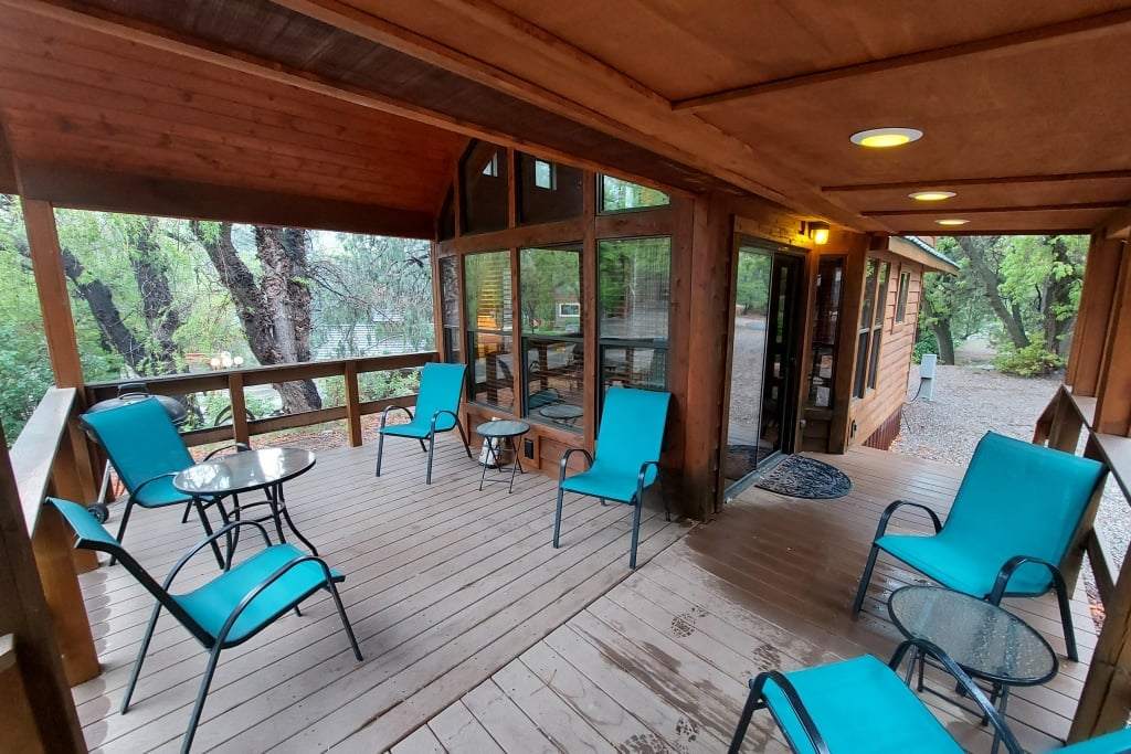Covered wooden porch with blue patio chairs and glass tables, attached to a cabin with large windows, surrounded by trees.