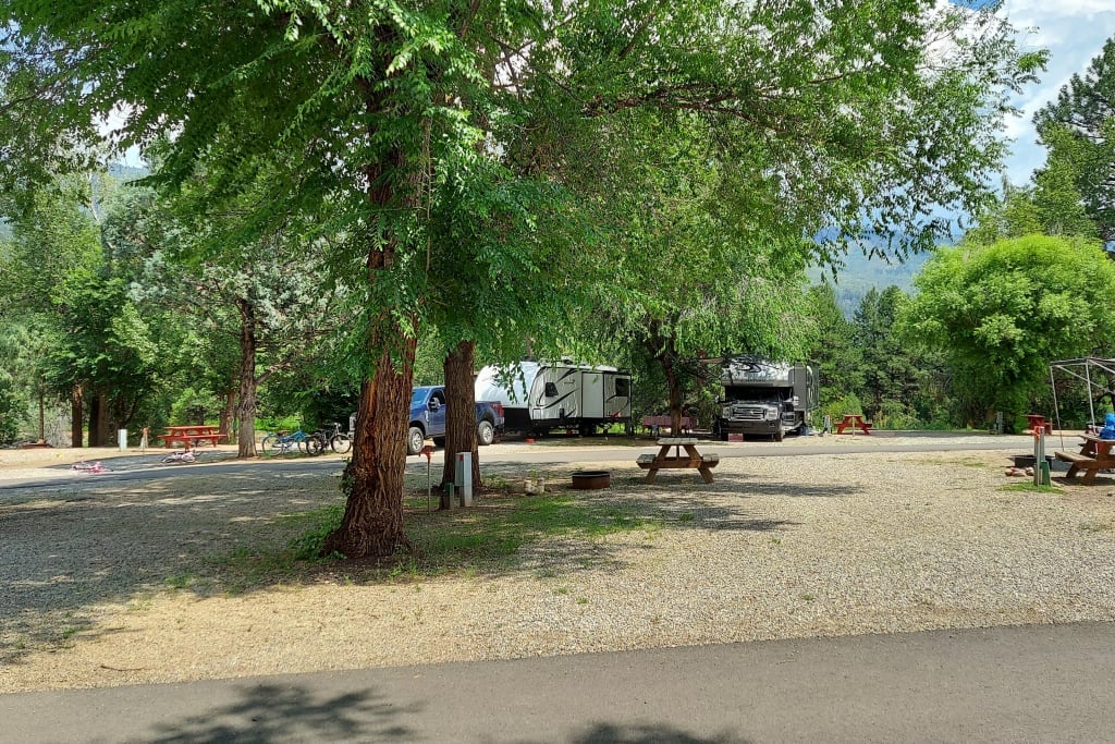 A gravel RV campsite with two RVs, picnic tables, trees, and scattered camping equipment under a sunny sky.