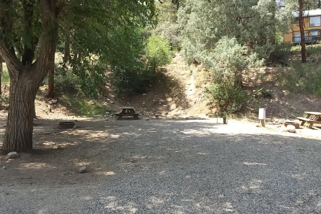 A gravel clearing with two picnic tables, a fire pit, and trees providing shade. A hillside with sparse vegetation rises in the background.