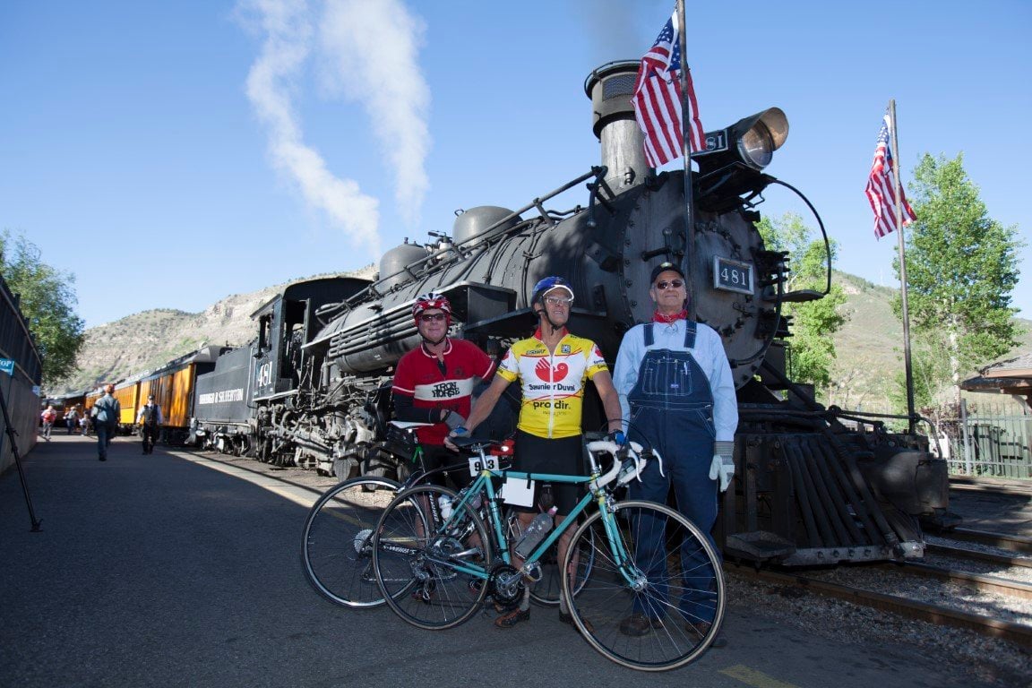 Two cyclists and a train engineer stand in front of a steam locomotive; two bicycles are parked in the foreground.