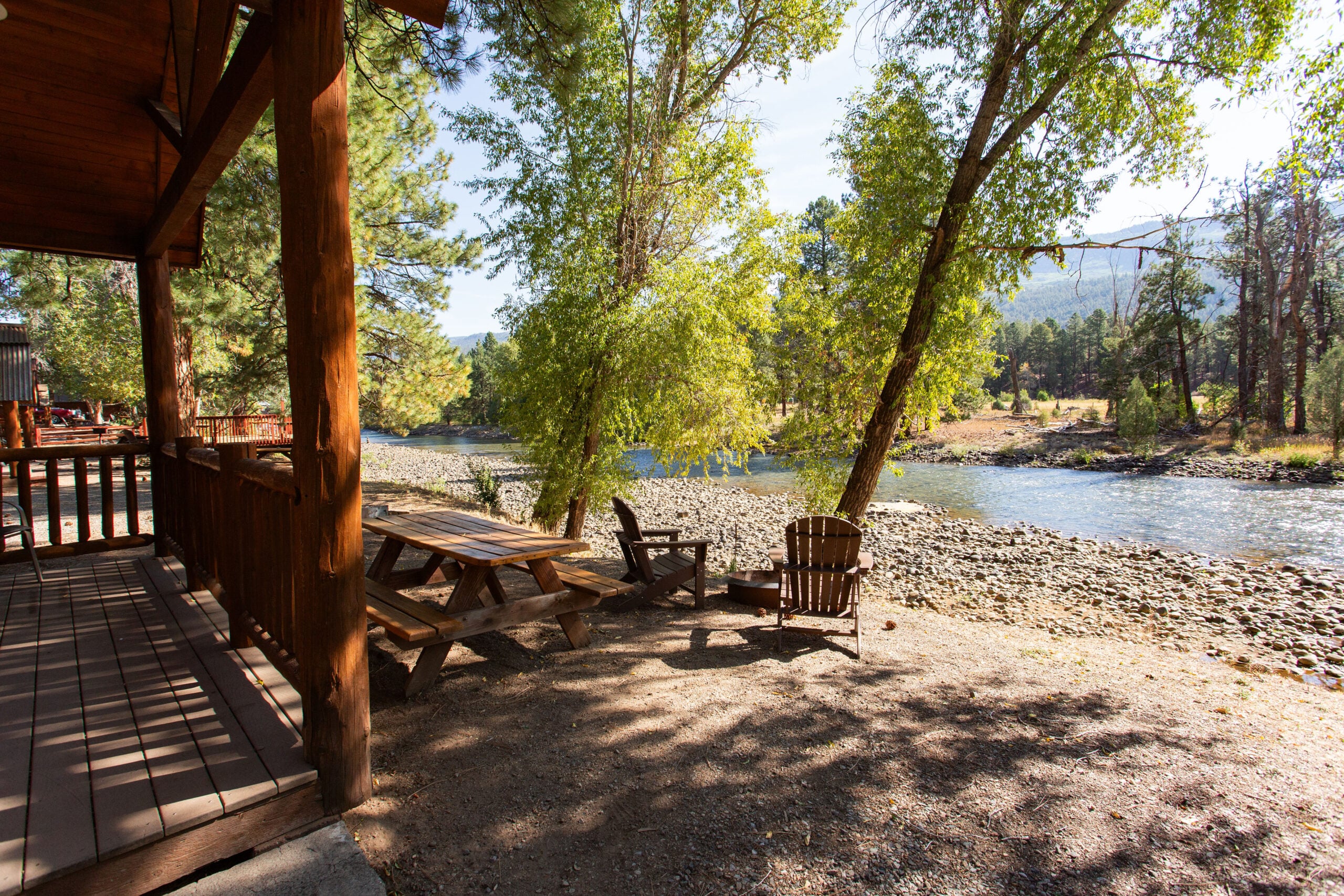 Wooden picnic table and Adirondack chairs sit beside a river, next to a cabin porch, surrounded by trees and sunlight.